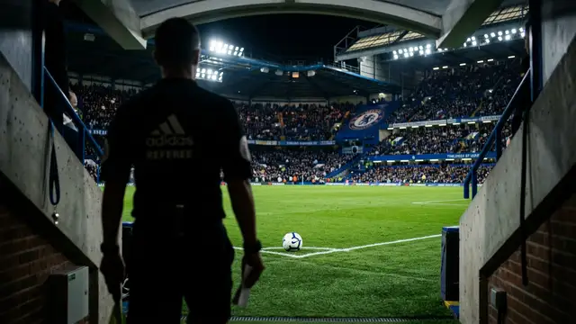 A referee's whistle and silhouette overlooking a brightly lit professional soccer pitch at night.