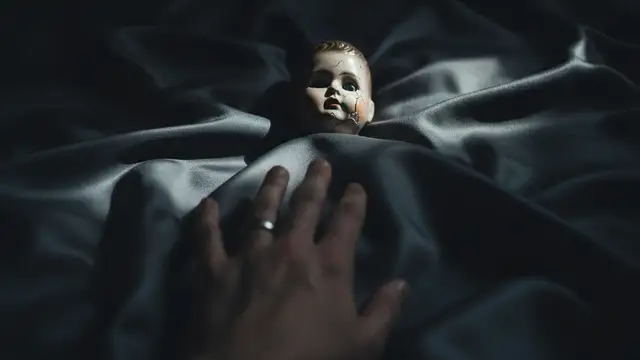 A porcelain doll on dark silk, a blurred hand with a ring reaching towards it.