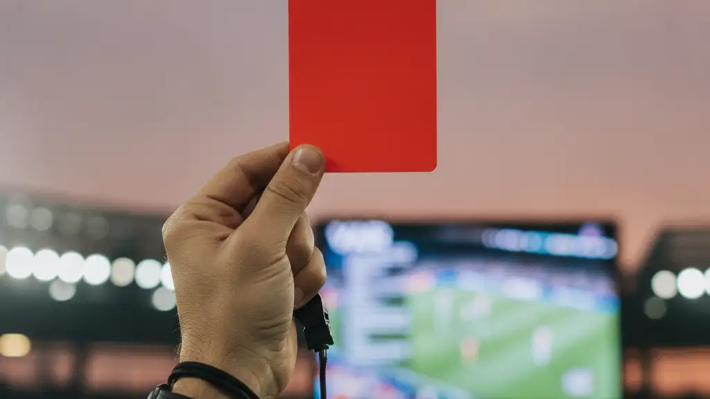 Referee's hand holding a red card against a blurred stadium background with VAR screen.