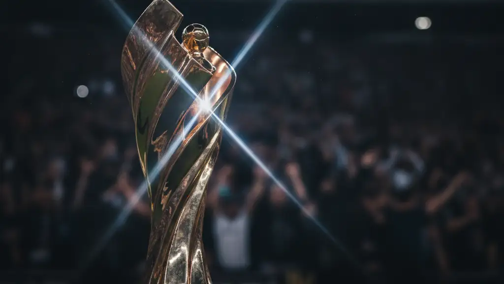 A golden football trophy reflecting stadium lights, with a blurred crowd in the background.