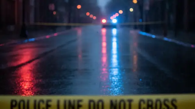 Emergency lights reflecting on a wet street with police tape in the foreground.