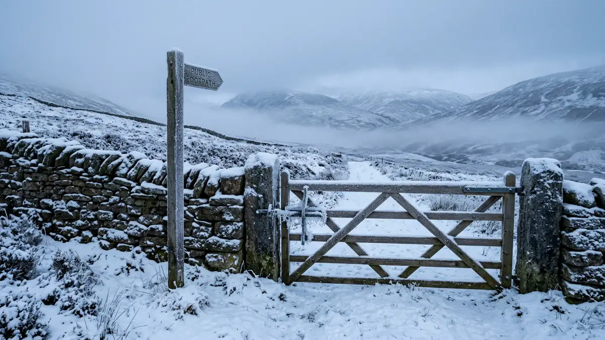 A snow-covered wooden gate and stone wall in a misty, frozen British rural landscape.