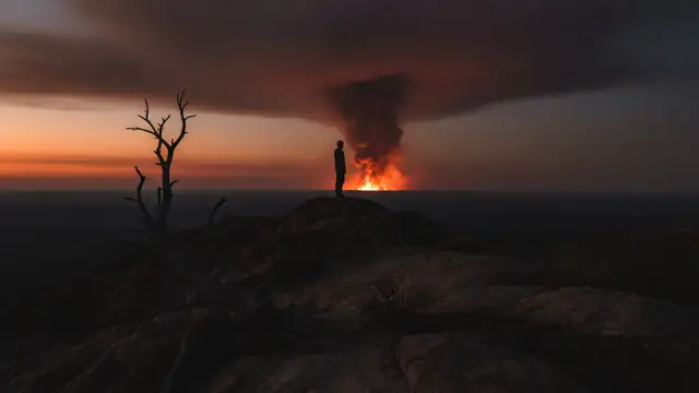 A lone silhouette on a ridge overlooks a distant, ominous orange and red smoke haze.
