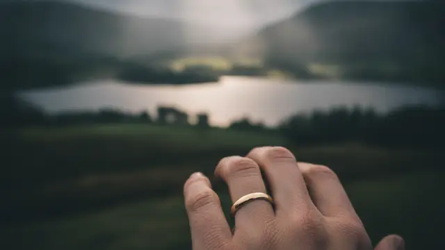 A hand with a wedding band, blurred Lake District landscape in background.