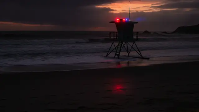 Emergency lights reflect on wet sand near a lifeguard tower at dusk.