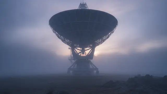 A massive radio telescope dish shrouded in atmospheric fog at twilight.