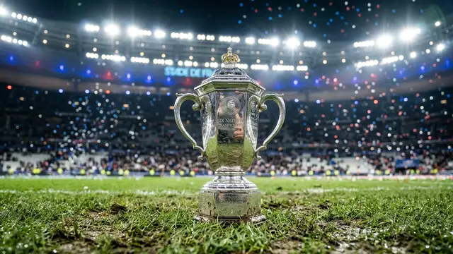 The Six Nations trophy sits on a rugby pitch under stadium lights with blue confetti.