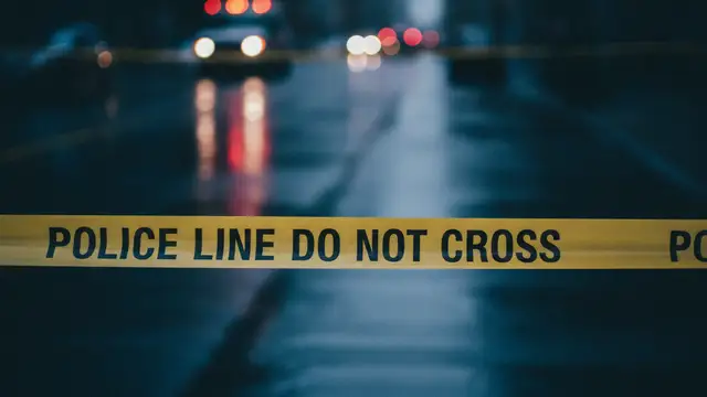 Yellow police tape in foreground, blurred red and blue emergency lights in background.