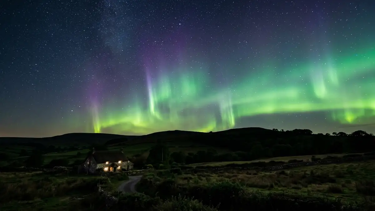 Green and purple northern lights shimmering in the night sky over a dark English landscape.