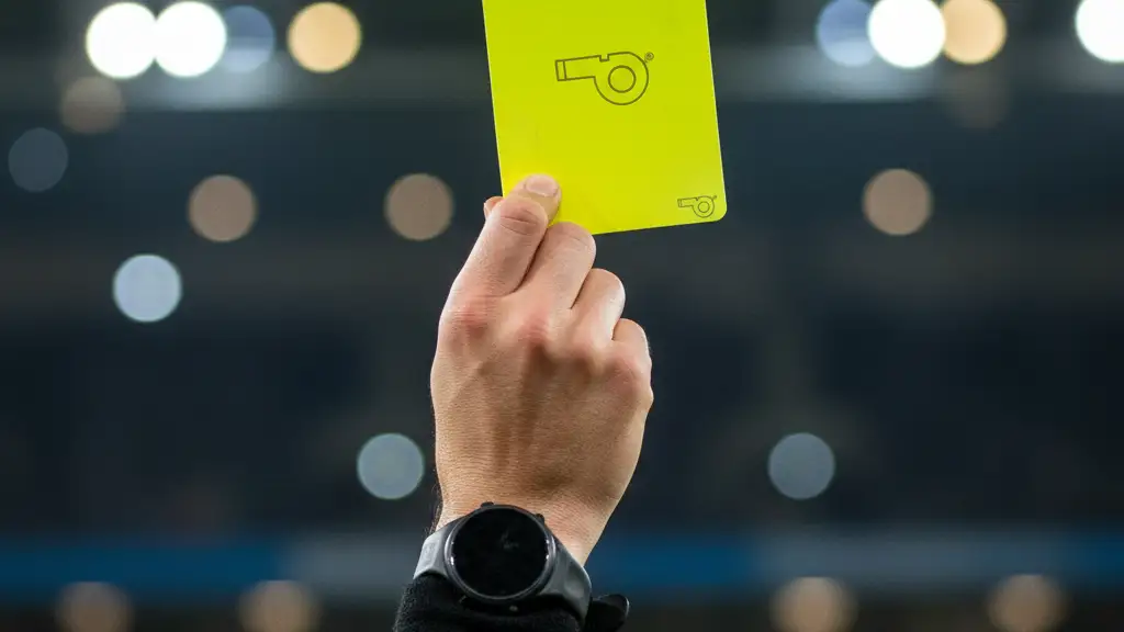 A referee's hand holds up a yellow card during a football match under stadium lights.