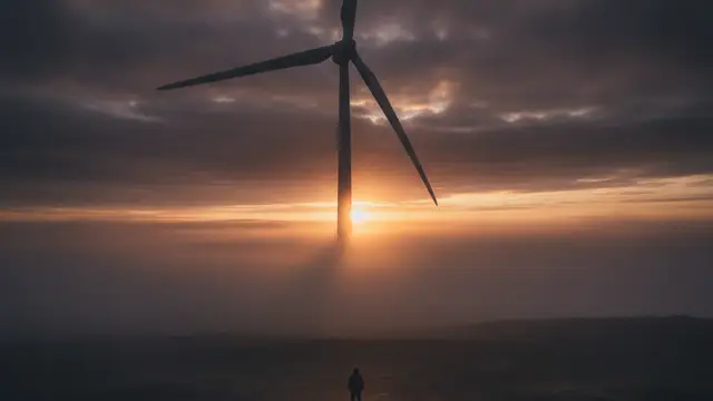 Giant wind turbine blade shrouded in mist at dawn, a figure looking up.