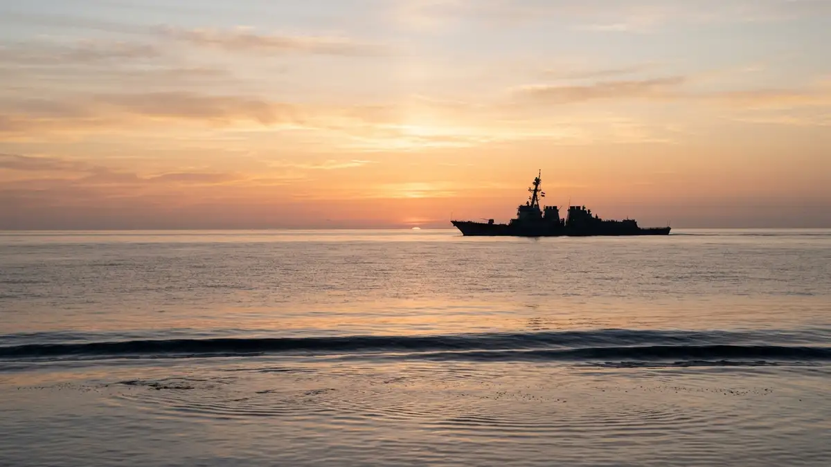 Naval destroyer silhouette on horizon at dawn, calm sea reflecting sunrise.
