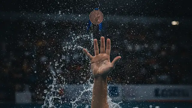 Swimmer's arm reaching for a bronze medal, face not visible, blurred stadium background.