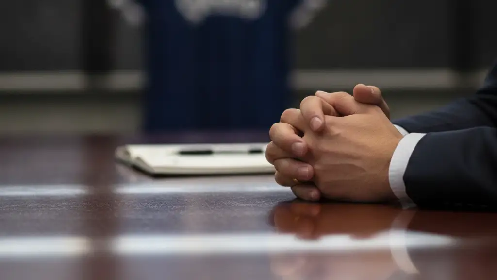 Manager's hands on a desk, stadium lights reflected, blurred football jersey.