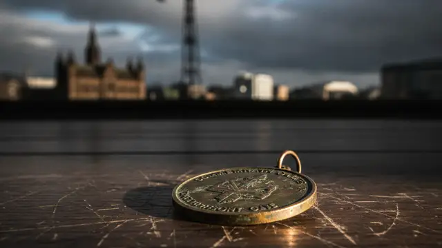 A tarnished Commonwealth Games medal on wood, blurred Glasgow architecture in background.
