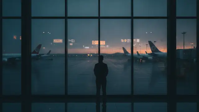 Silhouetted figure looking out airport window at rainy tarmac.
