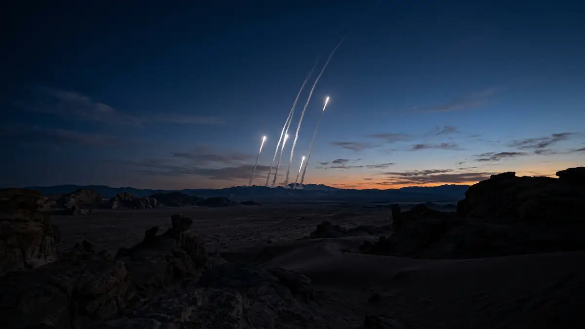 Long white vapor trails ascend into a dark desert sky at twilight.