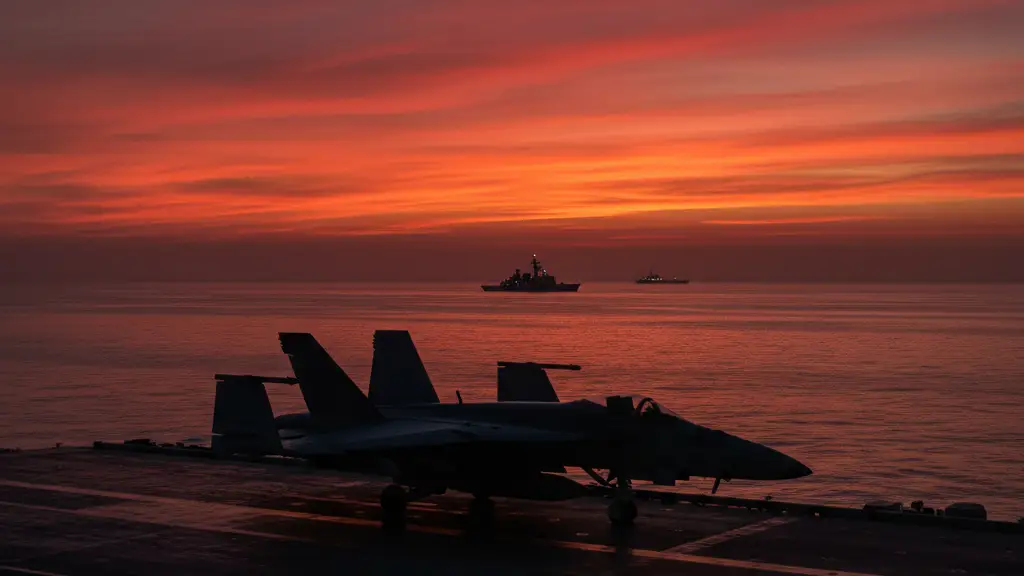 A military aircraft carrier deck at sunset with a silhouetted jet and naval escort.